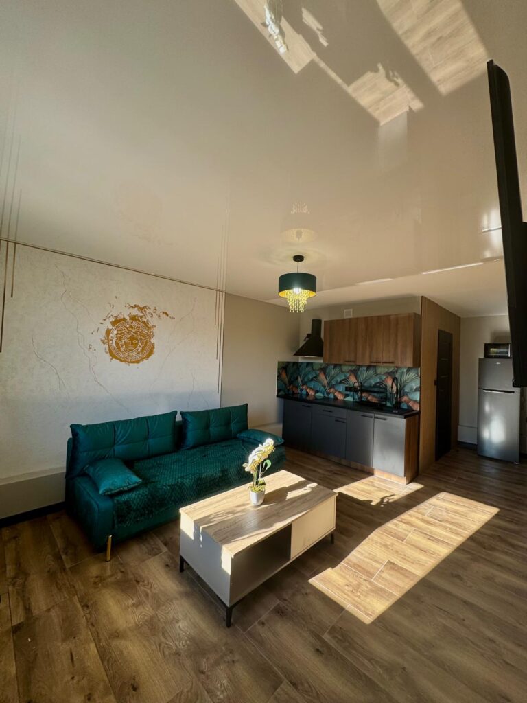 Nowoczesny salon z aneksem kuchennym – zielona kanapa, jasne drewniane podłogi i dekoracyjna ściana z motywem liści w kuchni