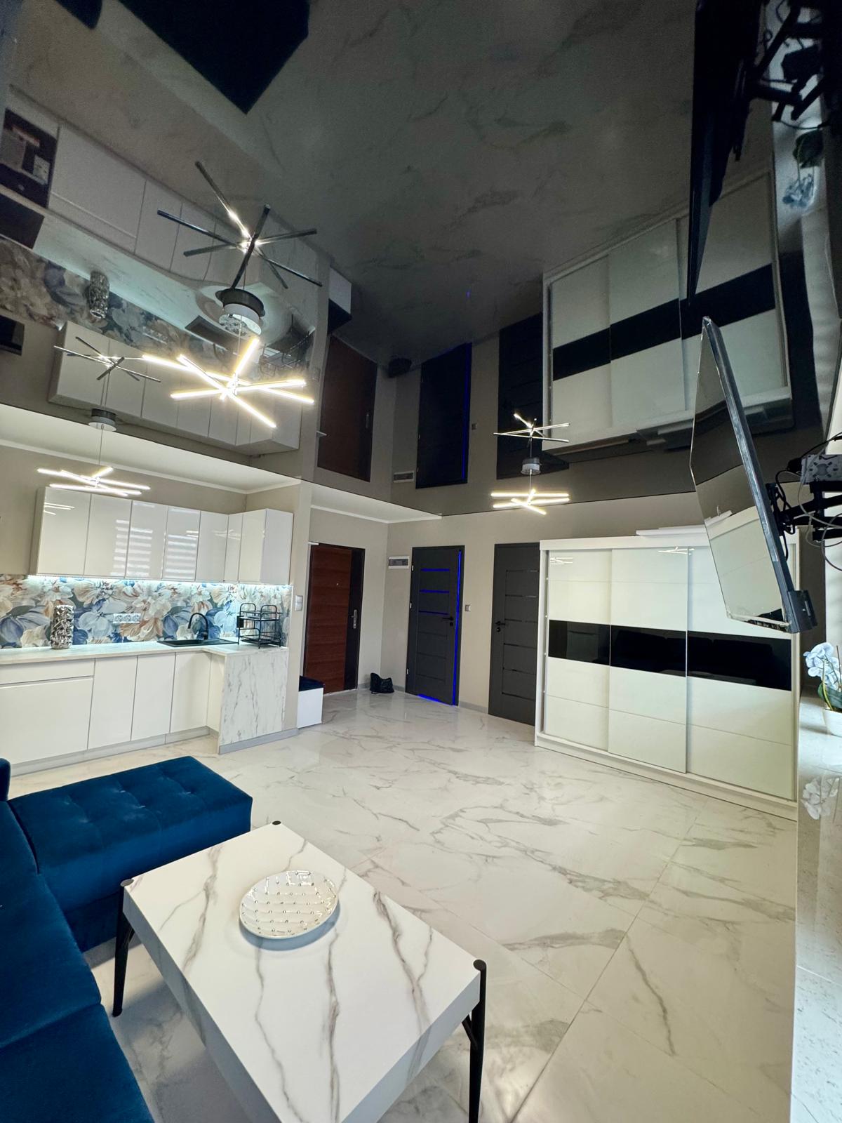 Eleganckie wnętrze salonu z białą, marmurową podłogą i lśniącym, czarnym sufitem napinanym, który optycznie powiększa przestrzeń. W pomieszczeniu znajduje się biała zabudowa kuchenna oraz szafa przesuwna.