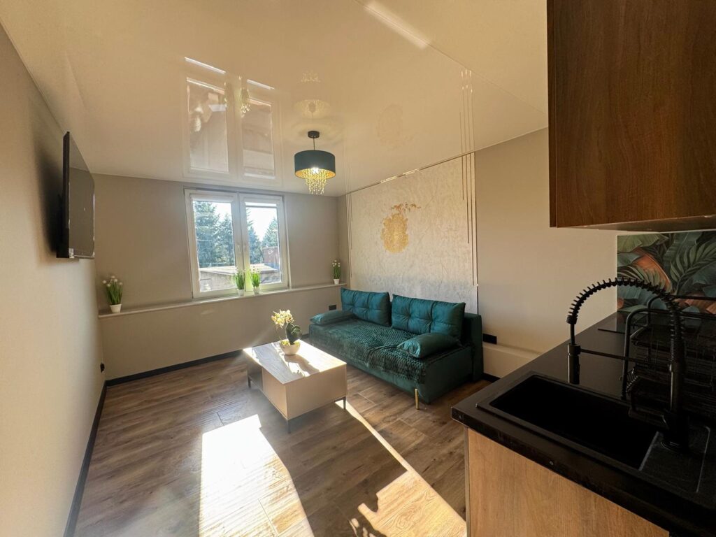 Nowoczesny salon z aneksem kuchennym – zielona kanapa, jasne drewniane podłogi i dekoracyjna ściana z logiem versauce