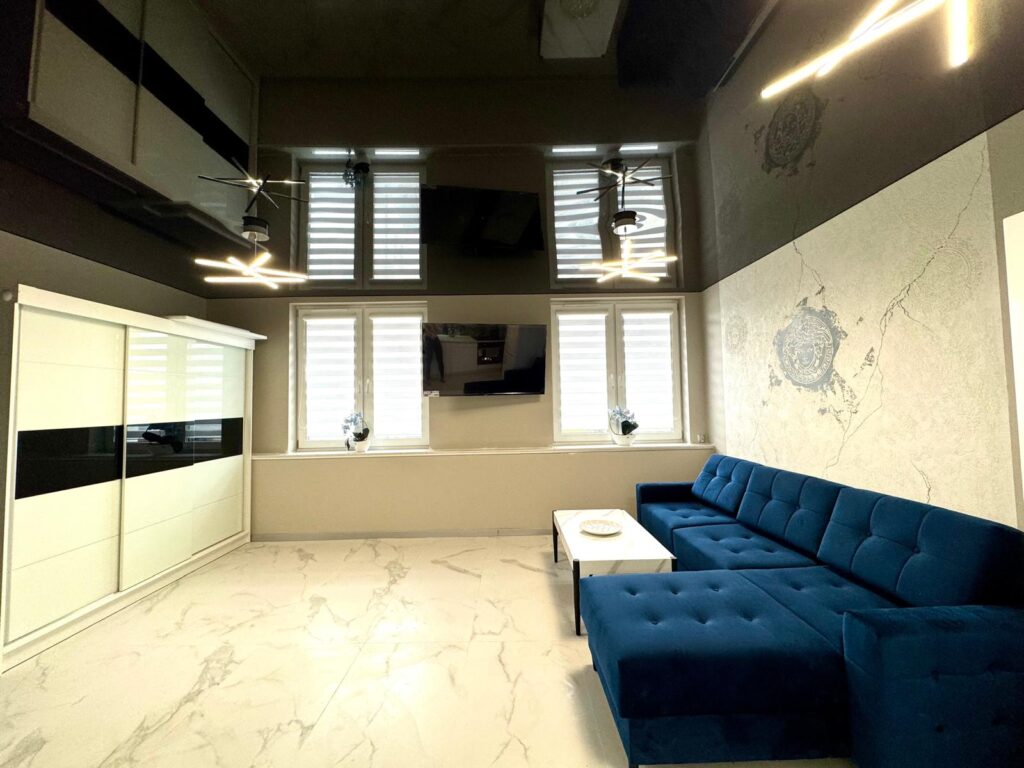 Eleganckie wnętrze salonu z białą, marmurową podłogą i lśniącym, czarnym sufitem napinanym, który optycznie powiększa przestrzeń. W pomieszczeniu znajduje się biała zabudowa kuchenna oraz szafa przesuwna.