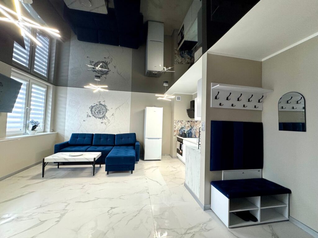 Eleganckie wnętrze salonu z białą, marmurową podłogą i lśniącym, czarnym sufitem napinanym, który optycznie powiększa przestrzeń. W pomieszczeniu znajduje się biała zabudowa kuchenna oraz szafa przesuwna.