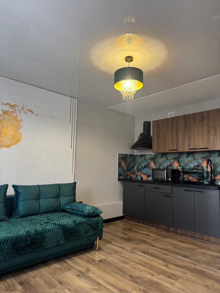 Nowoczesny salon z aneksem kuchennym – zielona kanapa, jasne drewniane podłogi i dekoracyjna ściana z motywem liści w kuchni