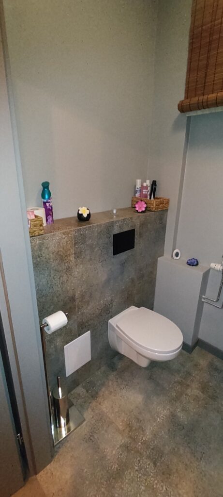 Nowoczesna toaleta w stylu SPA z podwieszaną miską WC i zabudowanym stelażem. Wnętrze utrzymane w odcieniach szarości z kamiennymi płytkami, z dekoracyjną półką nad toaletą, na której stoją kosmetyki, świeczki i drobne ozdoby. Całość tworzy spokojną, estetyczną i relaksującą przestrzeń.