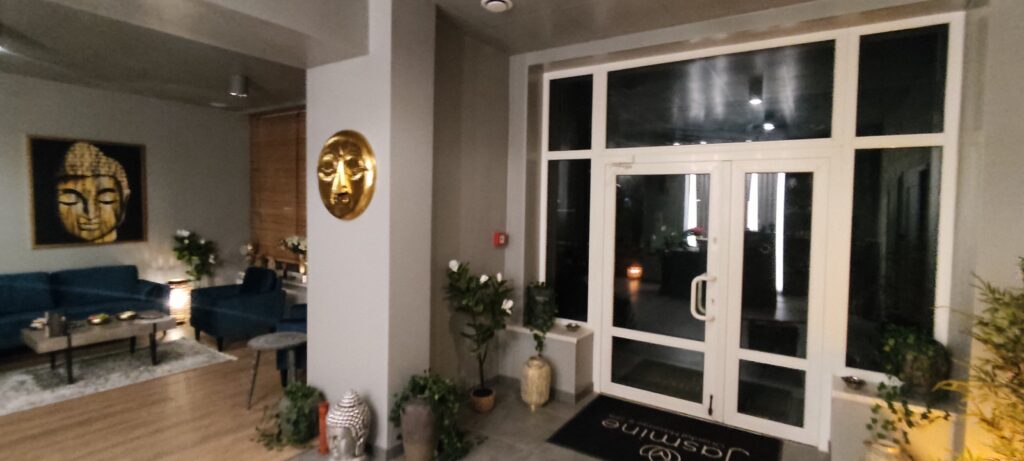 Strefa wejściowa do salonu SPA z przeszklonymi drzwiami i dużymi oknami. Po lewej stronie znajduje się elegancka poczekalnia z niebieskimi sofami, stolikiem kawowym i obrazem przedstawiającym twarz Buddy. Wnętrze utrzymane jest w spokojnej, relaksującej atmosferze, z dekoracjami w stylu orientalnym, roślinami oraz nastrojowym oświetleniem