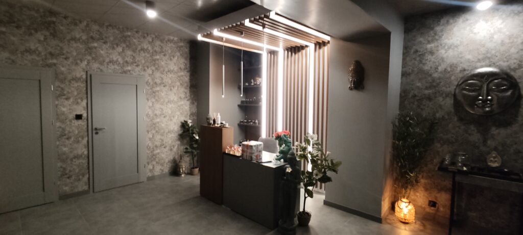 Eleganckie wnętrze strefy SPA z recepcją, utrzymane w stonowanych szarościach i ciepłych akcentach drewna. Centralnie znajduje się lada recepcyjna z dekoracjami i roślinami, nad którą wiszą nowoczesne lampy. W tle widoczne są półki z kosmetykami oraz podświetlona drewniana ścianka. Po prawej stronie znajduje się dekoracyjna rzeźba twarzy i nastrojowe oświetlenie, a po lewej drzwi prowadzące do innych pomieszczeń.