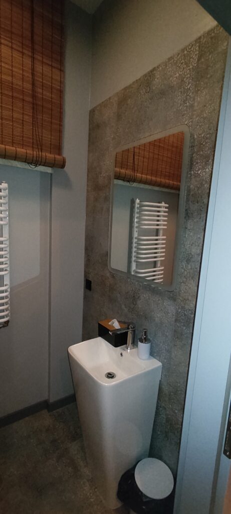 Mała, nowoczesna łazienka z umywalką stojącą przy ścianie, nad którą wisi prostokątne lustro. Ściany wykończone są szarymi płytkami o kamiennej fakturze, a na oknie znajduje się bambusowa roleta. Obok umywalki widać biały grzejnik drabinkowy oraz dozownik na mydło i pudełko na chusteczki.