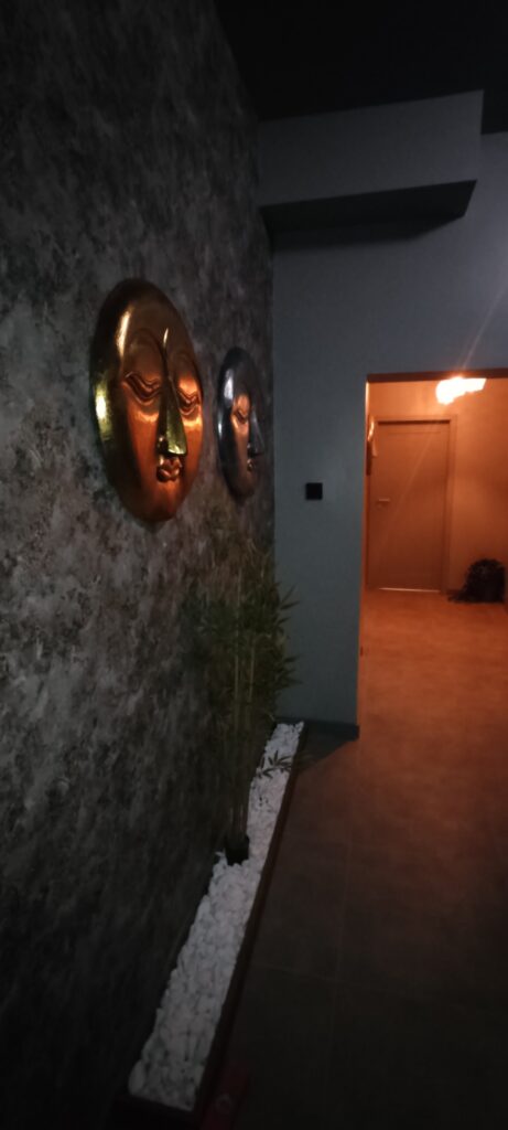 Ciemny, nastrojowy korytarz w stylu spa. Na ścianie po lewej stronie wiszą dwie dekoracyjne maski przedstawiające spokojne twarze w stylu orientalnym, w kolorach złotym i grafitowym. U dołu ściany znajduje się pas białych kamieni ozdobnych oraz roślina w donicy. Korytarz prowadzi do jasno oświetlonego przejścia z widocznymi drzwiami, a całość tworzy elegancką, relaksującą atmosferę.