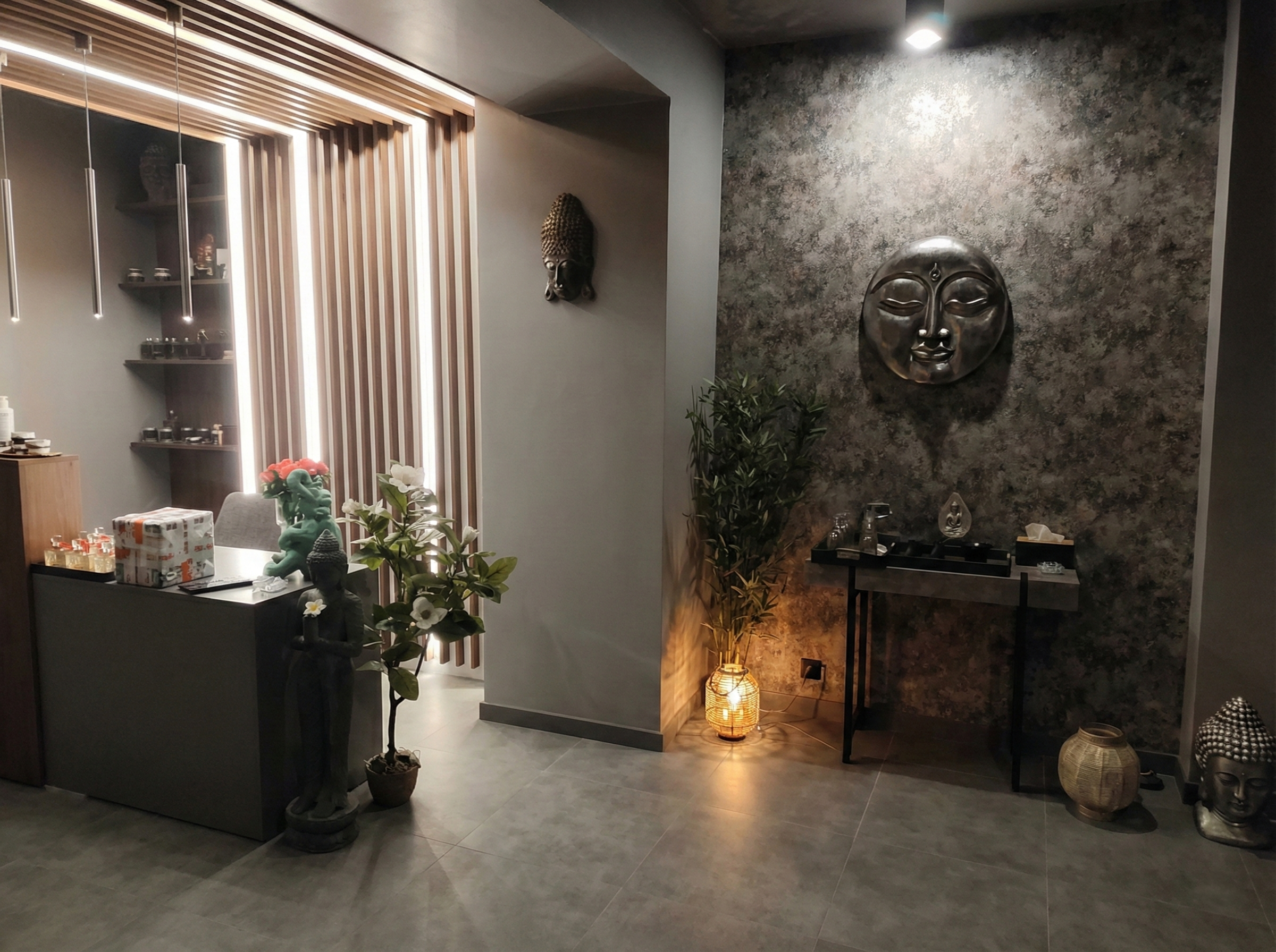 Recepcja salonu SPA z nowoczesną ladą, podświetlaną drewnianą ścianką i wiszącymi lampami. Po prawej stronie widoczna jest dekoracyjna ściana z rzeźbą twarzy w stylu orientalnym, stolik z akcesoriami oraz nastrojowe oświetlenie. Wnętrze utrzymane w ciemnych, eleganckich tonacjach szarości i drewna, z roślinami podkreślającymi relaksujący charakter miejsca.