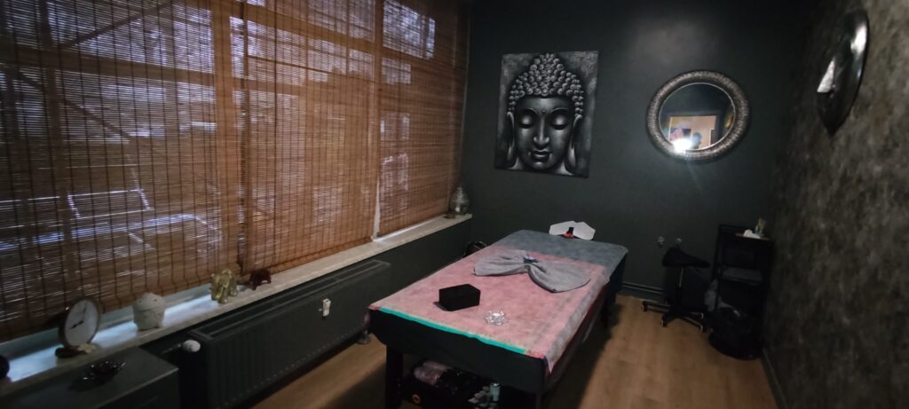 Przytulny gabinet masażu lub spa w ciemnej, stonowanej kolorystyce. Pośrodku stoi łóżko do masażu przykryte ręcznikiem. Na ścianie widoczny jest obraz przedstawiający twarz Buddy oraz okrągłe lustro. Po lewej stronie znajduje się duże okno z bambusowymi roletami, a na parapecie ustawione są małe figurki dekoracyjne. Wnętrze o spokojnej, relaksującej atmosferze, z drewnianą podłogą i subtelnym oświetleniem.