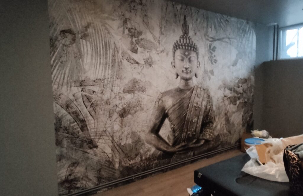 Wnętrze pomieszczenia w trakcie remontu. Na jednej ze ścian znajduje się duży, monochromatyczny mural przedstawiający medytującą postać Buddy w spokojnej pozie. Obok widać wysokie, wąskie okno przy suficie. Na podłodze leżą kartony, torby i materiały remontowe, co sugeruje, że przestrzeń nie jest jeszcze urządzona. Całość utrzymana w stonowanych, szaro-beżowych barwach.