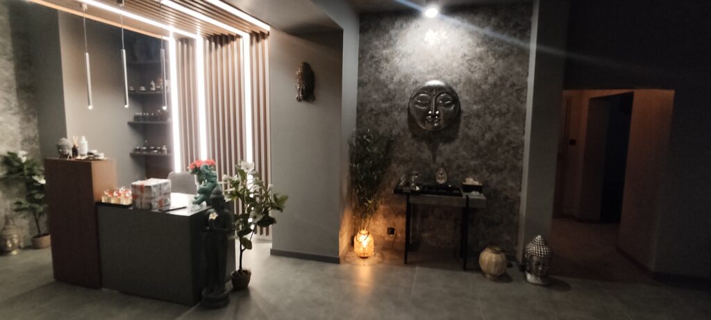 Recepcja salonu SPA z nowoczesną ladą, podświetlaną drewnianą ścianką i wiszącymi lampami. Po prawej stronie widoczna jest dekoracyjna ściana z rzeźbą twarzy w stylu orientalnym, stolik z akcesoriami oraz nastrojowe oświetlenie. Wnętrze utrzymane w ciemnych, eleganckich tonacjach szarości i drewna, z roślinami podkreślającymi relaksujący charakter miejsca.