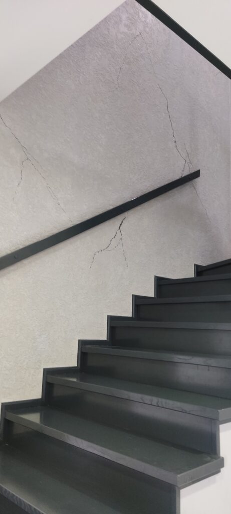 Schody w kolorze czarnym na tle jasnej ściany z dekoracyjnym tynkiem imitującym naturalne spękania.