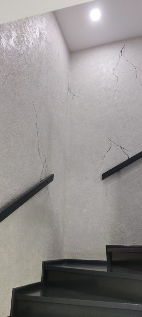 Klatka schodowa z dekoracyjnym tynkiem strukturalnym w jasnym kolorze z czarnymi żyłkami, czarne schody i oświetlenie sufitowe.