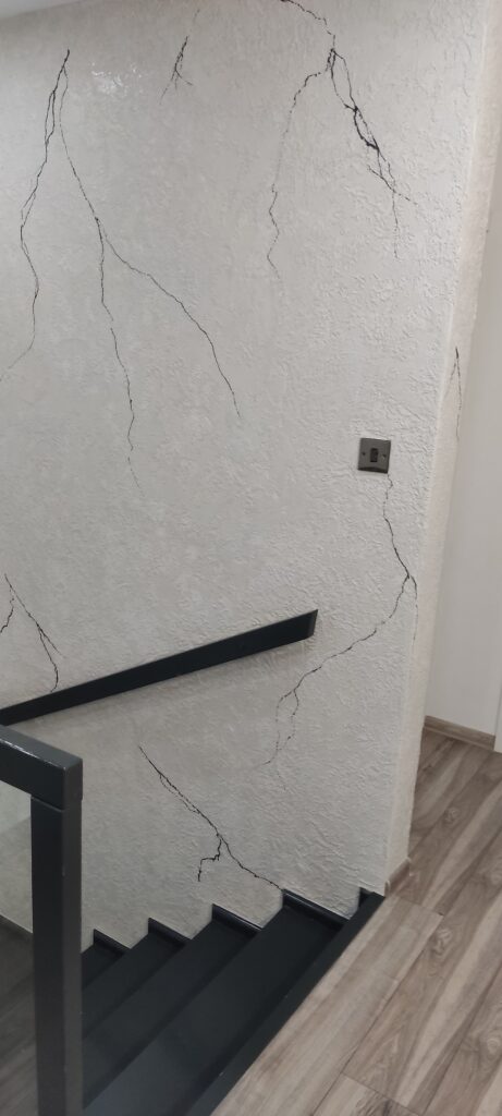 Ściana klatki schodowej wykończona jasnym tynkiem dekoracyjnym z efektem spękań i czarną poręczą.