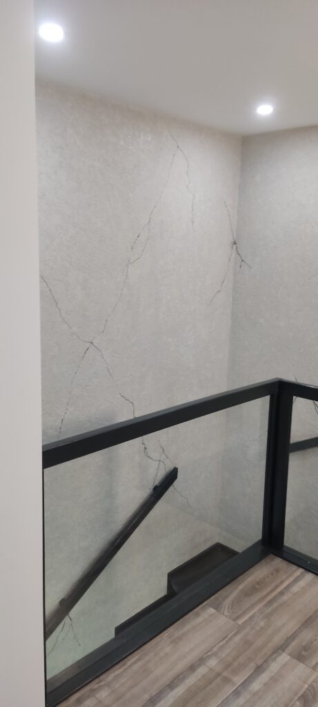 Nowoczesna klatka schodowa z przeszkloną balustradą, czarnymi elementami i dekoracyjnym tynkiem strukturalnym.