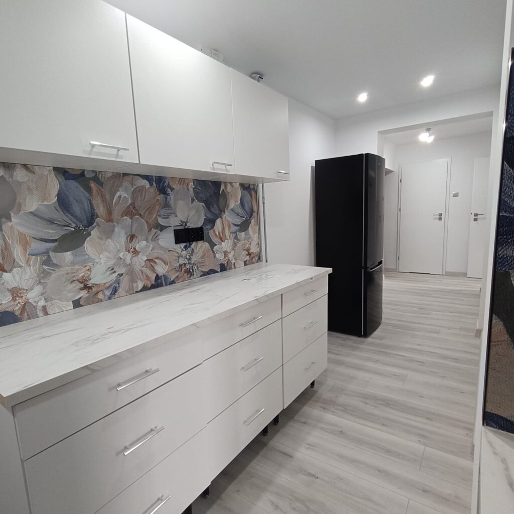 Nowoczesna kuchnia po remoncie z białą zabudową, czarną lodówką i dekoracyjną ścianą z motywem kwiatowym.